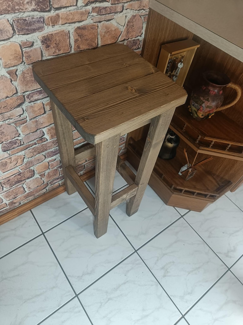 K&ouml;nnte beinhalten: Rustikaler Barhocker aus Holz mit quadratischer Sitzfl&auml;che und vier stabilen Beinen. Der Hocker ist aus hellbraunem Holz gefertigt, das die nat&uuml;rliche Maserung zeigt. Das Design ist schlicht und funktional, geeignet f&uuml;r K&uuml;che oder Bar.