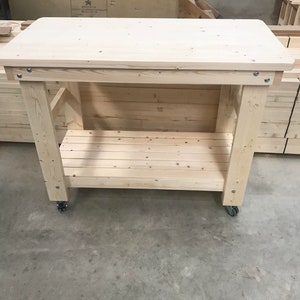 May include: A natural wood workbench with a single shelf and wheels. The workbench is made of unfinished pine wood and has a simple design.