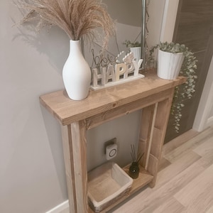 Op de afbeelding: Een rustieke houten console met een witte vaas met gedroogd pampasgras. Boven de tafel hangt een spiegel. Een kleine potplant en een decoratief bord met de tekst "Happy" staan ook op de tafel.