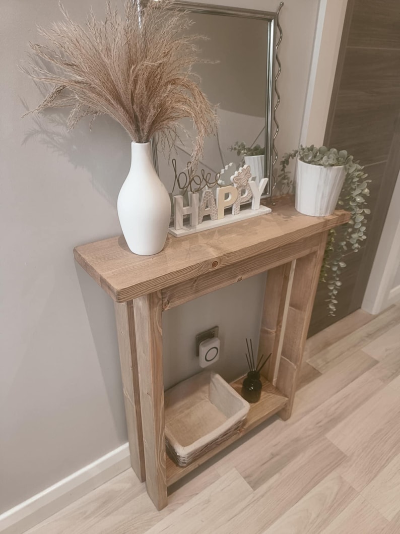 Op de afbeelding: Een rustieke houten console met een witte vaas met gedroogd pampasgras. Boven de tafel hangt een spiegel. Een kleine potplant en een decoratief bord met de tekst "Happy" staan ook op de tafel.