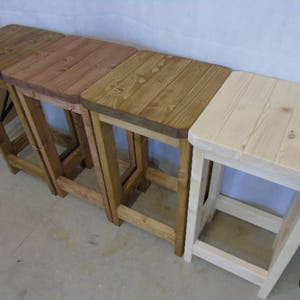 K&ouml;nnte beinhalten: Vier Holzst&uuml;hle in verschiedenen Braun- und Naturholzt&ouml;nen. Die Hocker haben quadratische Oberteile und abgewinkelte Beine, die eine rustikale &Auml;sthetik zeigen. Die Holzmaserung ist sichtbar und tr&auml;gt zum nat&uuml;rlichen Charme der M&ouml;bel bei.