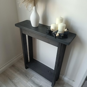 Op de afbeelding: Een donkergrijze houten consoletafel met een onderste plank. Een witte vaas met gedroogd siergras en drie witte kaarsen op een zwarte metalen houder staan op de tafel. De tafel staat tegen een witte muur.