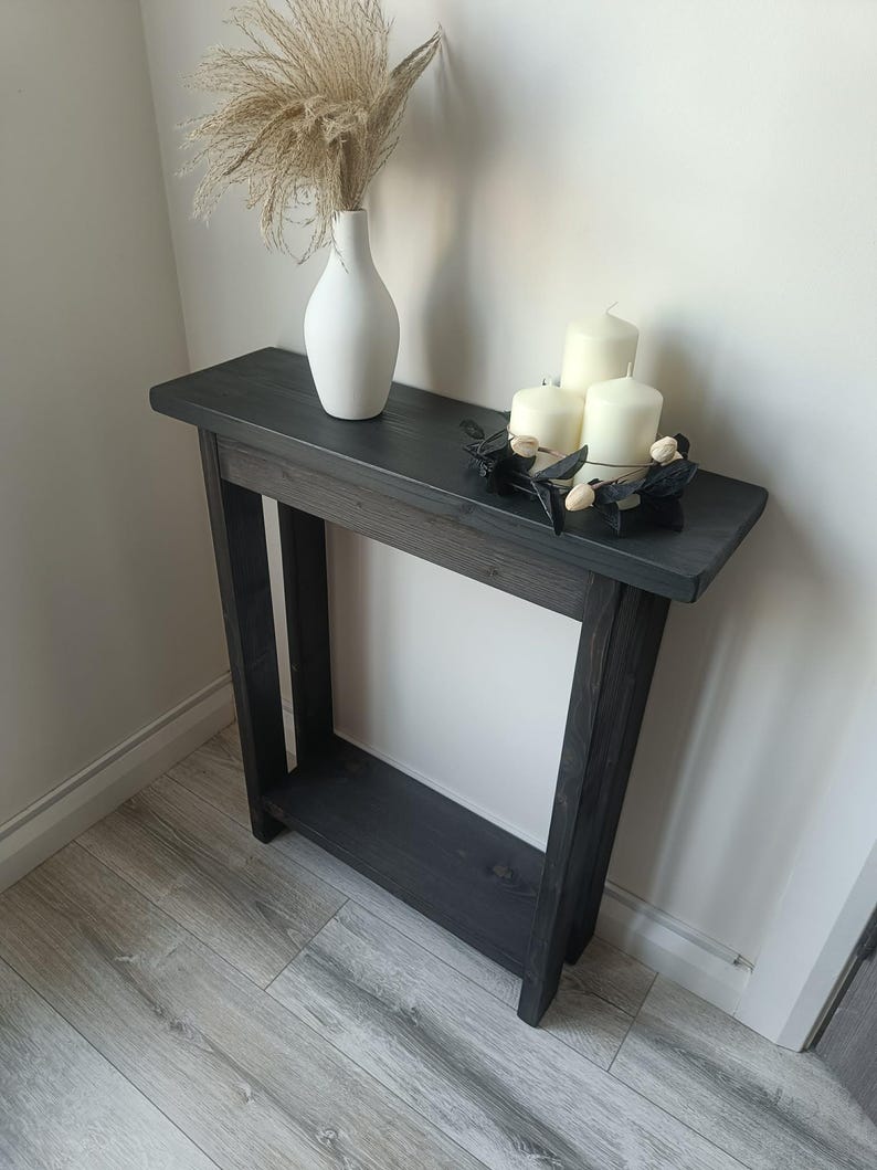 Op de afbeelding: Een donkergrijze houten consoletafel met een onderste plank. Een witte vaas met gedroogd siergras en drie witte kaarsen op een zwarte metalen houder staan op de tafel. De tafel staat tegen een witte muur.