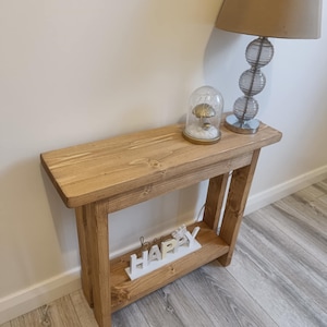 Op de afbeelding: Een lichtbruine houten consoletafel met een onderste plank. Op de tafel staan een glazen lamp met een beige kap en een decoratieve glazen koepel met een paddenstoel erin. Het woord "happy" staat op de onderste plank.