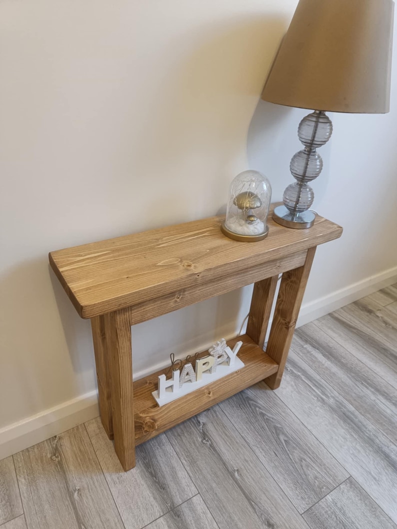 Op de afbeelding: Een lichtbruine houten consoletafel met een onderste plank. Op de tafel staan een glazen lamp met een beige kap en een decoratieve glazen koepel met een paddenstoel erin. Het woord "happy" staat op de onderste plank.