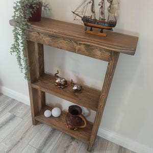May include: A wooden console table with three shelves. A potted plant and a model sailing ship sit on the top shelf. The lower shelves display elephant figurines, spheres, and a ceramic jug. The wood has a warm, brown colour.