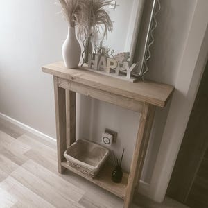Op de afbeelding: Een lichtgekleurde houten console met een plank, met een gevlochten mand, een zwarte diffuser en een witte vaas met gedroogde bloemen. Een decoratieve spiegel en een "bee happy" bord staan erop.
