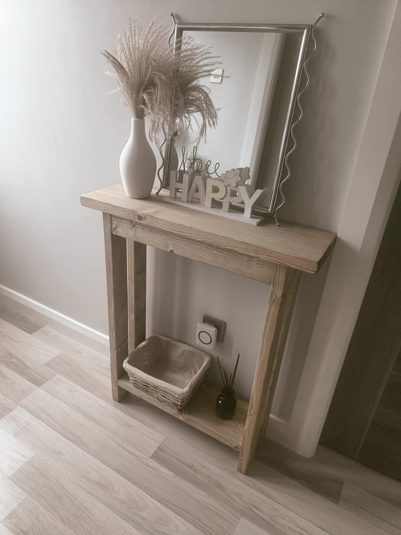 Op de afbeelding: Een lichtgekleurde houten console met een plank, met een gevlochten mand, een zwarte diffuser en een witte vaas met gedroogde bloemen. Een decoratieve spiegel en een "bee happy" bord staan erop.