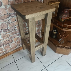 K&ouml;nnte beinhalten: Ein rustikaler Barhocker aus Holz mit quadratischer Sitzfl&auml;che und vier stabilen Beinen. Der Hocker ist aus hellbraunem Holz gefertigt und weist eine sichtbare Holzmaserung auf. Das Design ist schlicht und funktional, geeignet f&uuml;r K&uuml;che oder Bar.