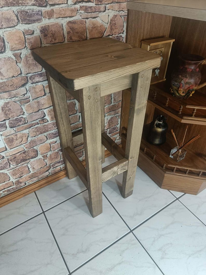 K&ouml;nnte beinhalten: Ein rustikaler Barhocker aus Holz mit quadratischer Sitzfl&auml;che und vier stabilen Beinen. Der Hocker ist aus hellbraunem Holz gefertigt und weist eine sichtbare Holzmaserung auf. Das Design ist schlicht und funktional, geeignet f&uuml;r K&uuml;che oder Bar.