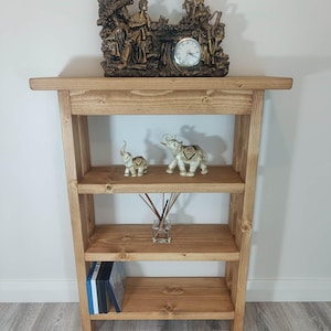 May include: A light brown wooden bookshelf with three shelves. The top shelf holds a decorative clock and a detailed sculpture. The middle shelf displays two elephant figurines and a glass vase with reeds. The bottom shelf has books.