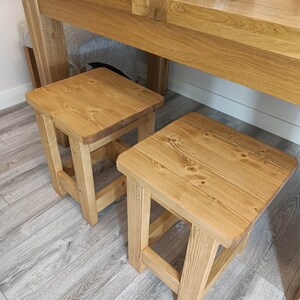 K&ouml;nnte beinhalten: Zwei kleine, quadratische Holzst&uuml;hle mit nat&uuml;rlichem Finish. Die Hocker sind aus hellfarbigem Holz gefertigt und haben ein schlichtes, robustes Design. Sie sind vor einem gro&szlig;en Holztisch platziert, was auf einen Ess- oder K&uuml;chenbereich hindeutet.
