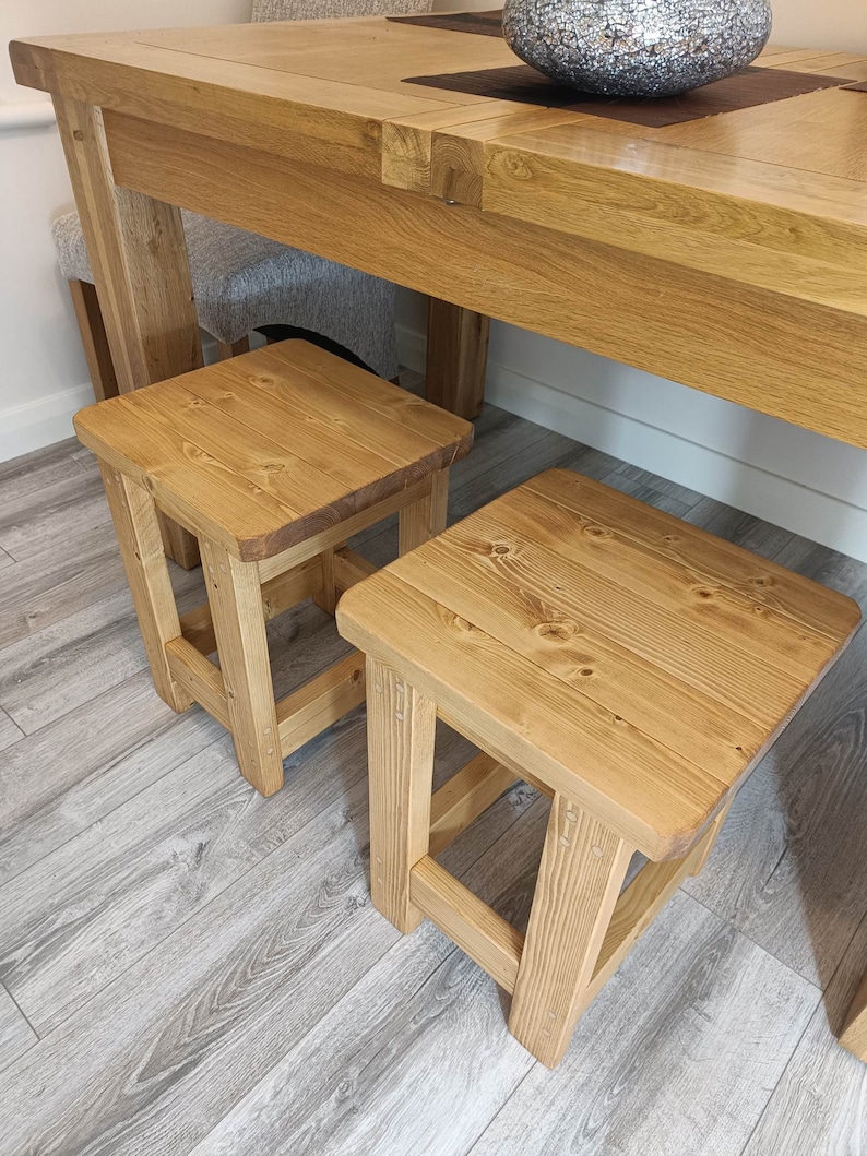 K&ouml;nnte beinhalten: Zwei kleine, quadratische Holzst&uuml;hle mit nat&uuml;rlichem Finish. Die Hocker sind aus hellfarbigem Holz gefertigt und haben ein schlichtes, robustes Design. Sie sind vor einem gro&szlig;en Holztisch platziert, was auf einen Ess- oder K&uuml;chenbereich hindeutet.