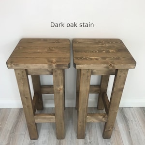 K&ouml;nnte beinhalten: Zwei Barhocker aus Holz mit dunkler Eichenbeize. Jeder Hocker hat eine quadratische Sitzfl&auml;che und vier stabile Beine. Der Text "Dark oak stain" steht &uuml;ber den Hockern.