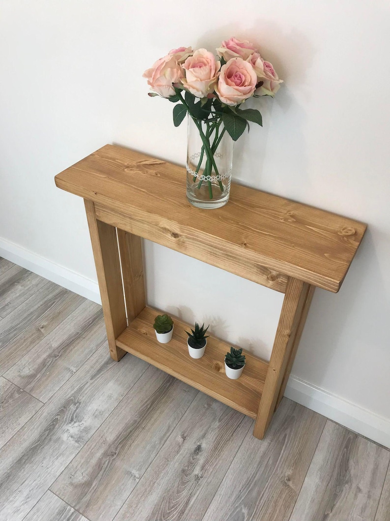 Op de afbeelding: Een lichtgekleurde houten console met een onderste plank. Een glazen vaas met roze rozen staat bovenop. Drie kleine vetplanten in potten staan op de onderste plank. De tafel staat tegen een witte muur.