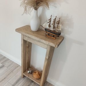 Op de afbeelding: Een rustieke houten console in lichtbruin. Bovenop staat een witte vaas met gedroogd siergras en een model zeilboot. De tafel heeft een kleine plank met decoratieve items. De console is gemaakt van natuurlijk hout.