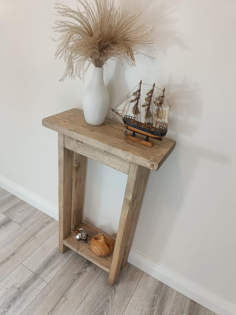 Op de afbeelding: Een rustieke houten console in lichtbruin. Bovenop staat een witte vaas met gedroogd siergras en een model zeilboot. De tafel heeft een kleine plank met decoratieve items. De console is gemaakt van natuurlijk hout.