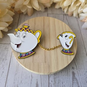 May include: A two-piece enamel pin set featuring a white teapot character with a purple and yellow lid and base, connected by a gold-toned metal chain to a smaller white teacup character with a yellow handle and a purple and yellow base.