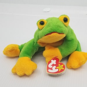 Puede incluir: Un beanie baby verde de rana con pies amarillos y una etiqueta de corazón rojo que dice "Beanie Babies" y "Baby".