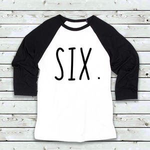 Pode incluir: Uma camisa branca e preta estilo raglan com a palavra "SIX" em letras grandes e pretas. A camisa tem mangas pretas e um corpo branco. O texto está centralizado na frente da camisa.