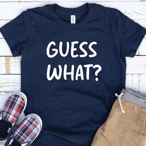May include: Navy blue t-shirt with the text "GUESS WHAT?" in white. The shirt is paired with khaki-colored pants and plaid slip-on shoes. The items are arranged on a white wooden surface.