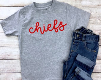 May include: A gray t-shirt with the word "chiefs" written in red cursive lettering.