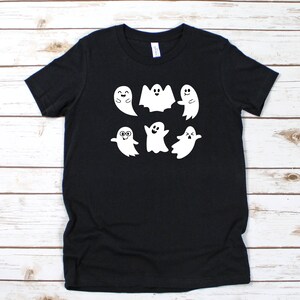 May include: Black t-shirt featuring a white graphic of six cartoon ghosts with various expressions. The ghosts are smiling, waving, and wearing glasses. The shirt is made of a soft material and is suitable for Halloween or everyday wear.