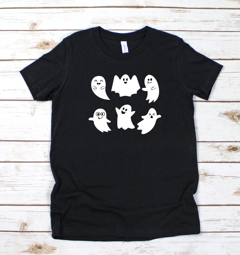May include: Black t-shirt featuring a white graphic of six cartoon ghosts with various expressions. The ghosts are smiling, waving, and wearing glasses. The shirt is made of a soft material and is suitable for Halloween or everyday wear.