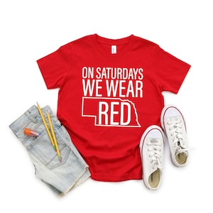 Op de afbeelding: Rood T-shirt met de tekst "On Saturdays We Wear Red" en een omtrek van de staat Nebraska. Het shirt ligt plat op een witte achtergrond met een paar witte sneakers en een blauwe spijkerbroek.