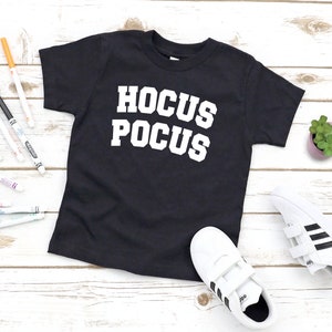 May include: Black t-shirt with the words "HOCUS POCUS" in large, white, block letters. The shirt is displayed on a light wood-grain surface, with white sneakers and colorful markers nearby. A small potted plant is also visible.