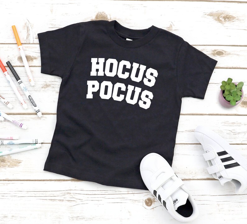 May include: Black t-shirt with the words "HOCUS POCUS" in large, white, block letters. The shirt is displayed on a light wood-grain surface, with white sneakers and colorful markers nearby. A small potted plant is also visible.