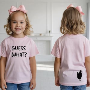 以下が含まれることがあります： 「GUESS WHAT?」の黒い文字が入った薄いピンク色のTシャツ。Tシャツの背面には、ニワトリの黒いシルエットがあります。子供は青いジーンズとピンクのリボンを身に着けています。