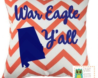 May include: Square decorative pillow with a white and orange chevron pattern. The pillow features the text "War Eagle y'all" in blue script, along with a blue outline of the state of Alabama.
