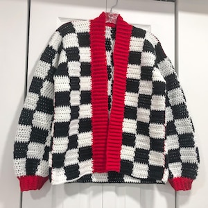 May include: A handmade crochet cardigan in a black and white checkerboard pattern. The open front and cuffs are trimmed with red. The sweater is hanging on a clear hanger against a white background.