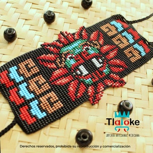 May include: A black beaded bracelet with a colourful Aztec design. The bracelet features red, blue, green, and white beads, with the word "Tloke" in the bottom right corner. The bracelet is on a woven surface, with black beads scattered around.