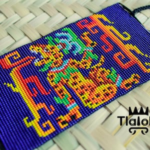 Puede incluir: Una pulsera de cuentas con un diseño vibrante que presenta una figura similar a un jaguar y patrones geométricos en tonos de azul, naranja, amarillo y verde. La pulsera tiene un fondo azul oscuro y cordones negros. La marca "Tlaloke" es visible.