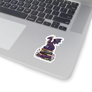May include: A die-cut sticker featuring a purple dragon with gold accents perched atop a stack of three vintage-style books. The dragon has intricate wing patterns, and the base of the book stack is surrounded by small pink and purple flowers and green leaves.