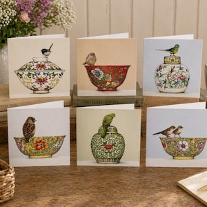 May include: A set of six square greeting cards with illustrations of birds on ornate bowls and vases. The cards display detailed artwork with bright colours and floral patterns, set against neutral backgrounds. The cards are arranged on a wooden surface.
