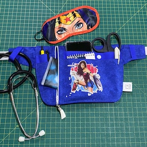 May include: A blue medical fanny pack featuring a Wonder Woman graphic, a black stethoscope, and a Wonder Woman eye mask. The fanny pack has pockets for pens, a phone, and medical tools. The eye mask is red, blue, and gold.