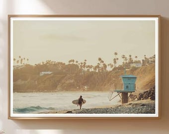 May include: Framed print of a beach scene with a surfer carrying a surfboard. A lifeguard tower stands on the right, and palm trees and a hillside are in the background. The image has a warm, golden color palette.