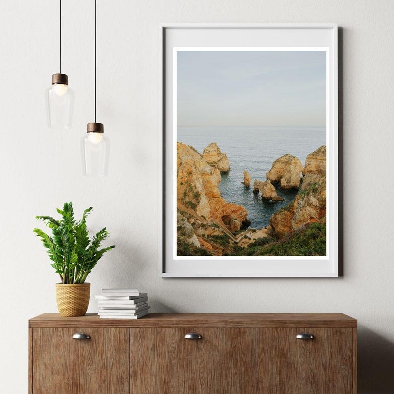 K&ouml;nnte beinhalten: Gerahmter Landschaftsdruck mit einer K&uuml;stenansicht mit goldbraunen Klippen und blauem Meer. Das Kunstwerk wird auf einem Holzschrank mit einer Topfpflanze und H&auml;ngeleuchten pr&auml;sentiert.