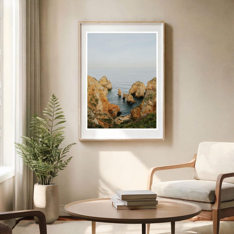 K&ouml;nnte beinhalten: Gerahmter Landschaftsdruck mit einer K&uuml;stenansicht. Das Bild zeigt zerkl&uuml;ftete, orangefarbene Klippen, die auf den blauen Ozean treffen, unter einem blassen Himmel. Der Druck wird in einem Raum mit neutralen T&ouml;nen zusammen mit einer Topfpflanze und einem runden Couchtisch ausgestellt.