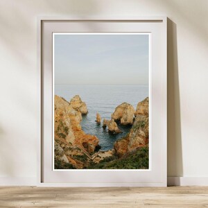 K&ouml;nnte beinhalten: Ein gerahmter Landschaftsdruck mit einer K&uuml;stenlandschaft. Das Bild zeigt goldbraune Felsformationen, die aus dem blauen Meer unter einem blassen Himmel aufragen. Ein Holzsteg ist links zu sehen, der zum Wasser f&uuml;hrt. Der Druck ist in einem wei&szlig;en Rahmen.