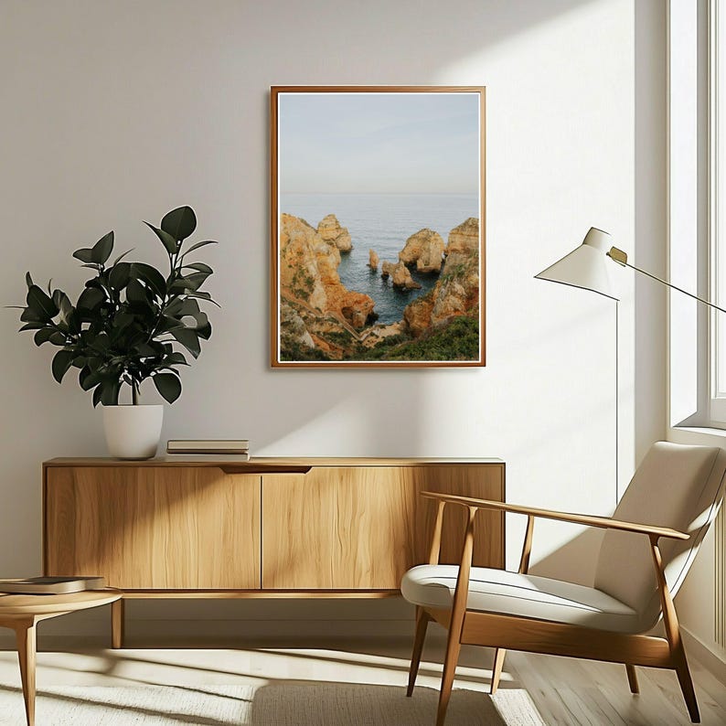 K&ouml;nnte beinhalten: Ein gerahmter Druck zeigt eine K&uuml;stenlandschaft mit Felsen und Meer. Der Druck hat einen Holzrahmen und h&auml;ngt an einer wei&szlig;en Wand &uuml;ber einem Holzschrank. Eine Topfpflanze und ein moderner Stuhl befinden sich ebenfalls im Raum.