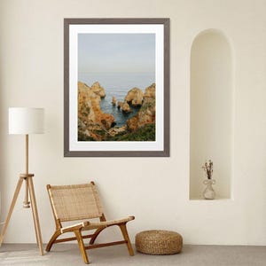 K&ouml;nnte beinhalten: Ein gerahmter Landschaftsdruck mit einer K&uuml;stenansicht mit orangefarbenen und braunen Felsformationen vor blauem Meer und Himmel. Der Druck befindet sich in einem grauen Rahmen und wird in einem Raum mit einem Holzstuhl und einer Stehlampe ausgestellt.