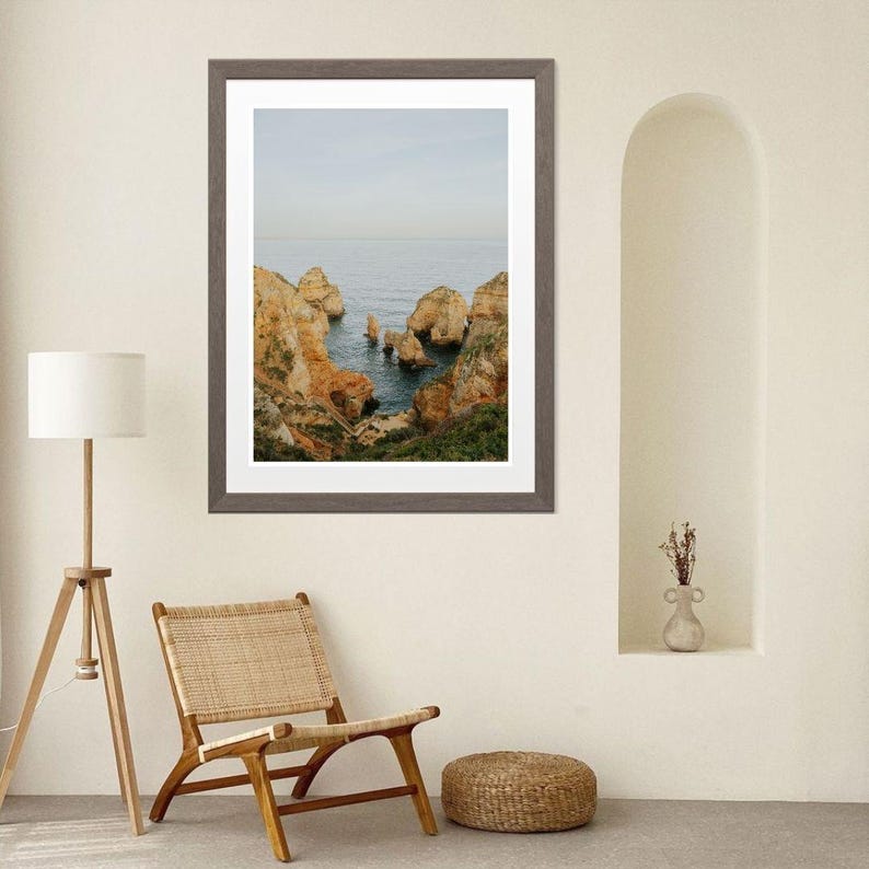 K&ouml;nnte beinhalten: Ein gerahmter Landschaftsdruck mit einer K&uuml;stenansicht mit orangefarbenen und braunen Felsformationen vor blauem Meer und Himmel. Der Druck befindet sich in einem grauen Rahmen und wird in einem Raum mit einem Holzstuhl und einer Stehlampe ausgestellt.
