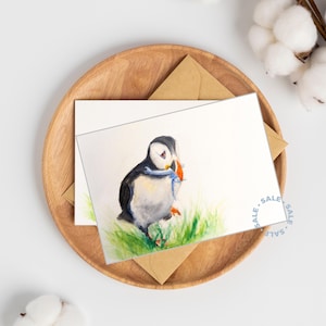 Könnte beinhalten: Aquarellzeichnung eines Papageientauchers mit Fisch im Schnabel, auf eine weiße Karte gedruckt. Die Karte liegt auf einem Holztablett mit einem braunen Umschlag. Der Vogel ist schwarz, weiß und orange, mit grünem Gras.