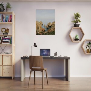K&ouml;nnte beinhalten: Ein gerahmter Druck einer K&uuml;stenlandschaft mit Felsen und Meer. Der Druck h&auml;ngt &uuml;ber einem Schreibtisch mit Laptop, Stuhl und Lampe. Ein B&uuml;cherregal und sechseckige Regale mit Pflanzen sind ebenfalls zu sehen.