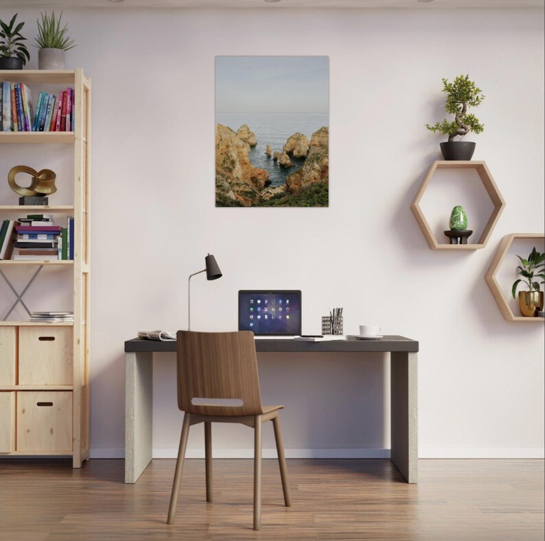 K&ouml;nnte beinhalten: Ein gerahmter Druck einer K&uuml;stenlandschaft mit Felsen und Meer. Der Druck h&auml;ngt &uuml;ber einem Schreibtisch mit Laptop, Stuhl und Lampe. Ein B&uuml;cherregal und sechseckige Regale mit Pflanzen sind ebenfalls zu sehen.
