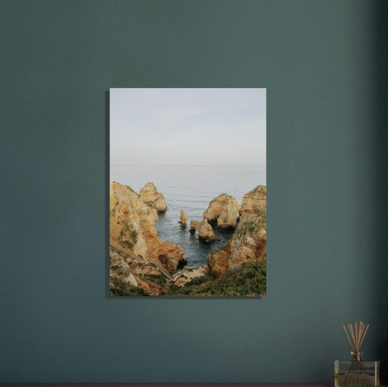 K&ouml;nnte beinhalten: Ein Landschaftsdruck mit einer K&uuml;stenlandschaft mit Felsformationen und dem Meer. Das Bild zeigt goldbraune Klippen und Felsnadeln vor einem ruhigen, blau-grauen Meer unter einem blassen Himmel. Das Kunstwerk wird an einer Wand ausgestellt.